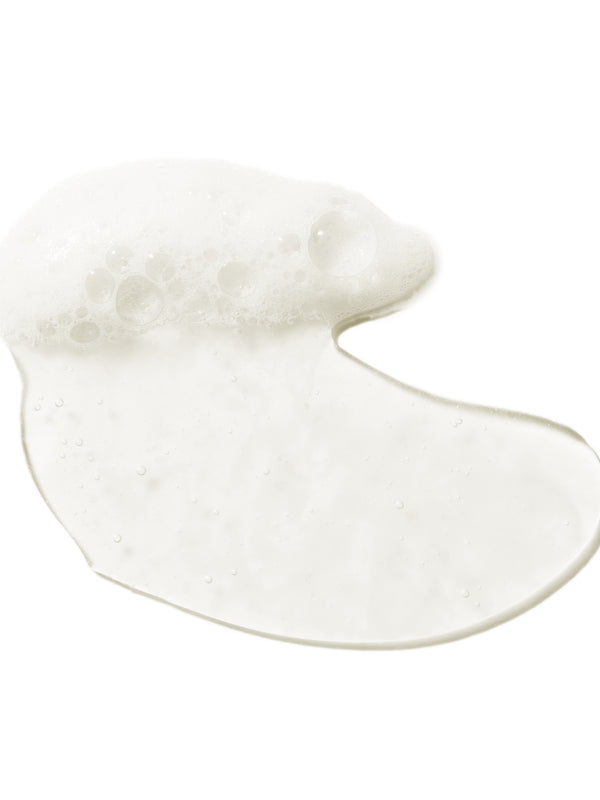 A glossy, translucent swatch of 100% Pure Glossy Locks Glossing Shampoo, forming an uneven puddle with white foam and small bubbles along one edge.