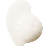 A pale, cloudy swatch of 100% Pure Glossy Locks Repair Shampoo, forming a glossy puddle with a patch of froth and small bubbles at the right.