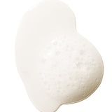 A pale, cloudy swatch of 100% Pure Glossy Locks Repair Shampoo, forming a glossy puddle with a patch of froth and small bubbles at the right.