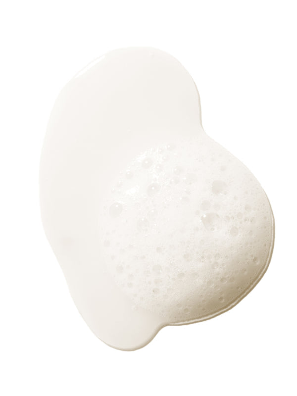 A pale, cloudy swatch of 100% Pure Glossy Locks Repair Shampoo, forming a glossy puddle with a patch of froth and small bubbles at the right.