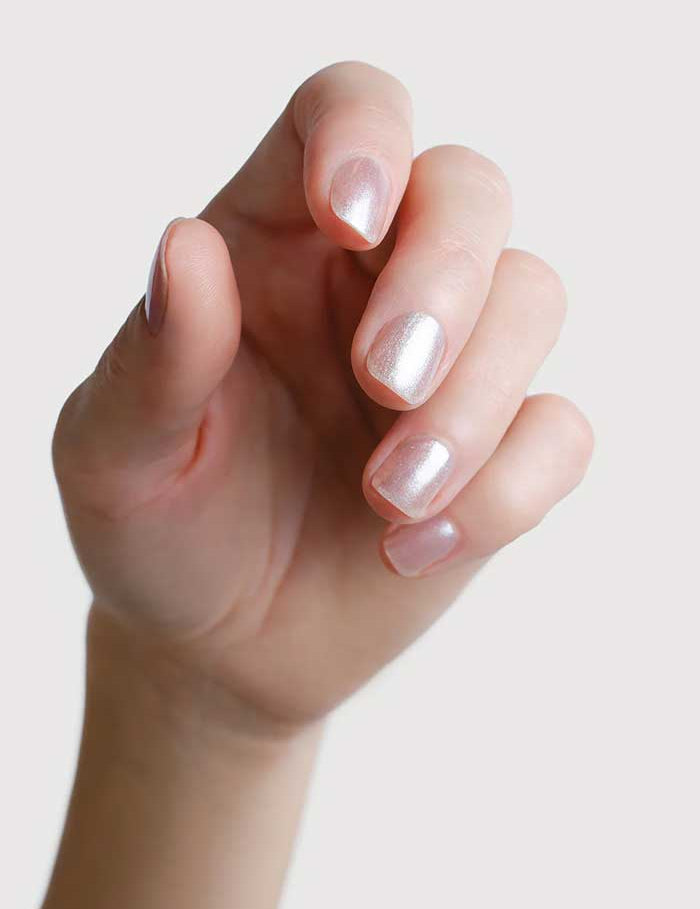 A close-up of gently curved fingers displays nails painted with Sienna Byron Bay Nail Polish - Glimmer 10ml, a shiny light pink alabaster shimmer, set against a plain white background.