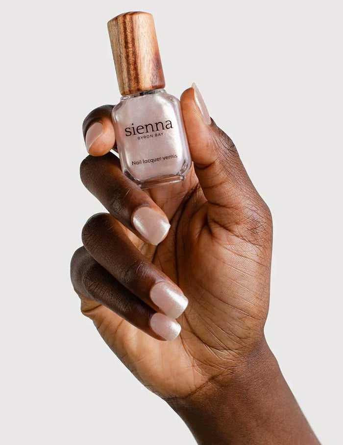 A hand with neatly manicured nails holds a 10ml bottle of Sienna Byron Bay Nail Polish in the shade Glimmer, featuring a wooden cap, against a plain white background.