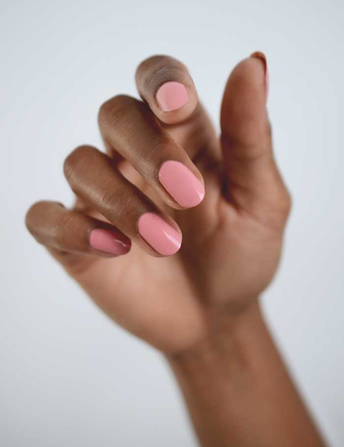 A close-up of a medium brown-skinned hand with neatly manicured nails painted in glossy light pink Sienna Byron Bay Nail Polish - Glowing 10ml, shown against a plain light background.