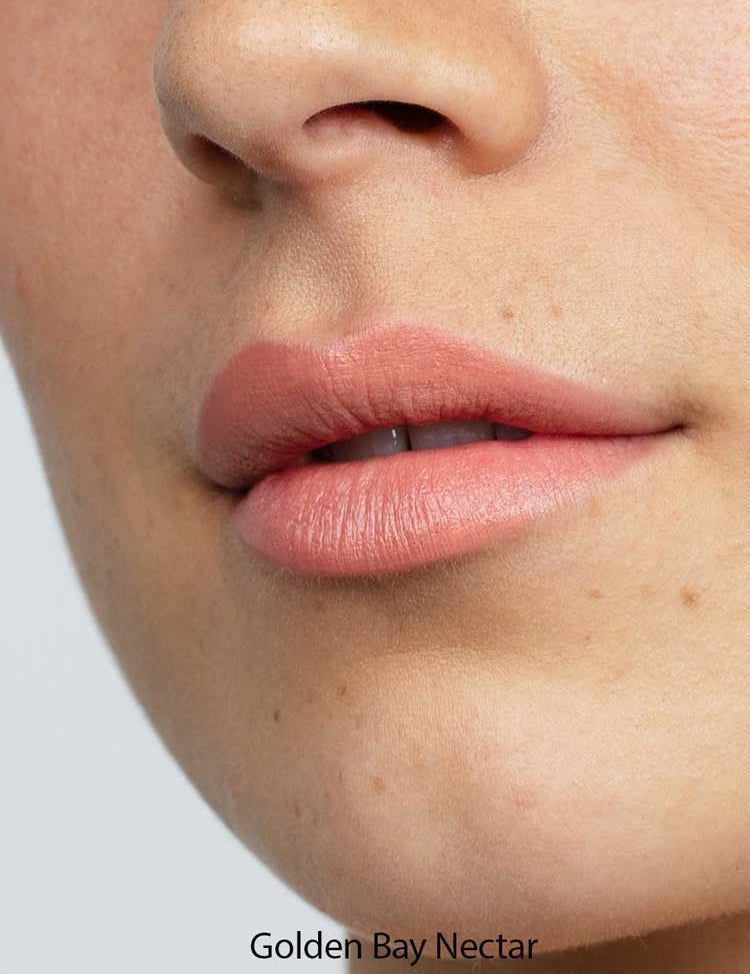 Close-up of lips in a matte, natural pink from Antipodes Moisture Boost Natural Lipsticks 4g. Skin looks smooth with freckles and light texture. "Golden Bay Nectar" at the bottom highlights long-lasting, natural color.