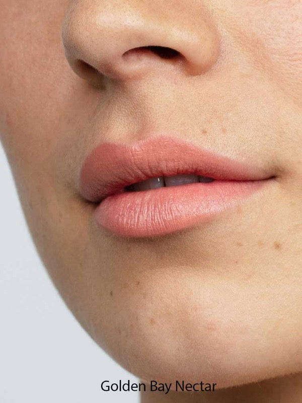 Close-up of lips in a matte, natural pink from Antipodes Moisture Boost Natural Lipsticks 4g. Skin looks smooth with freckles and light texture. "Golden Bay Nectar" at the bottom highlights long-lasting, natural color.