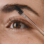 A close-up of a person’s eye and eyebrow, with a spoolie brushing the hairs—clearly defined by Madara Grow Fix Lash And Brow Booster Clear 4.25ml. The person has brown eyes, clear skin, and visible freckles.