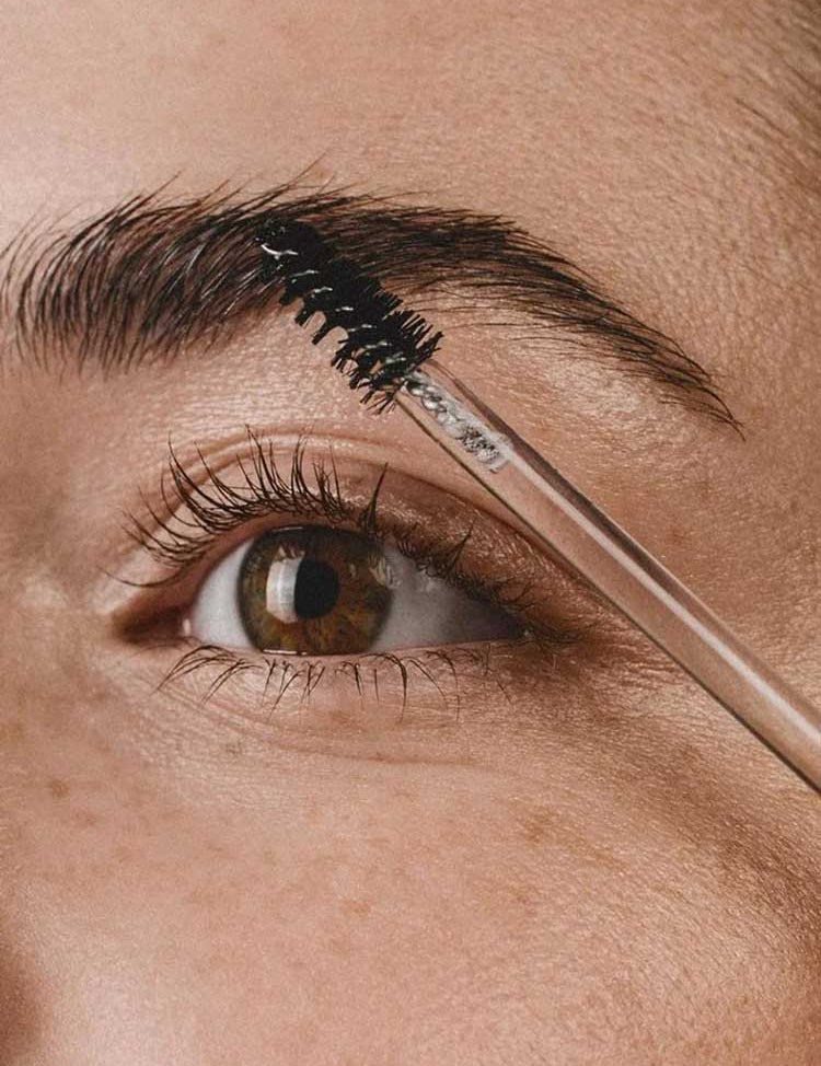 A close-up of a person’s eye and eyebrow, with a spoolie brushing the hairs—clearly defined by Madara Grow Fix Lash And Brow Booster Clear 4.25ml. The person has brown eyes, clear skin, and visible freckles.