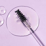 A close-up of the Madara Grow Fix Lash And Brow Booster Clear 4.25ml wand on a light purple surface, with clear gel in circular pools surrounding the brush tip.
