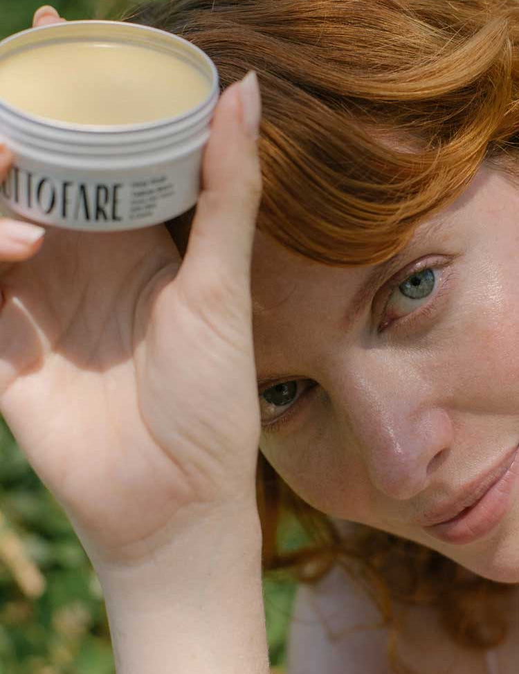 A red-haired person holds a round tin of Tuttofare Holy Grail Tallow Balm 70g close to their face, displaying it to the camera against a blurred background of greenery and sunlight.