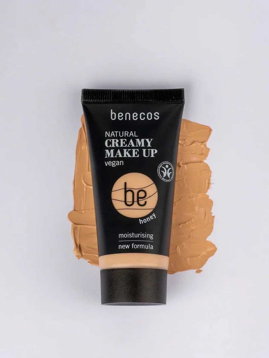 A tube of Benecos Natural Creamy Makeup 30ml in honey sits on a matching foundation smear. The packaging highlights its vegan, moisturising formula and new formula against a light background.
