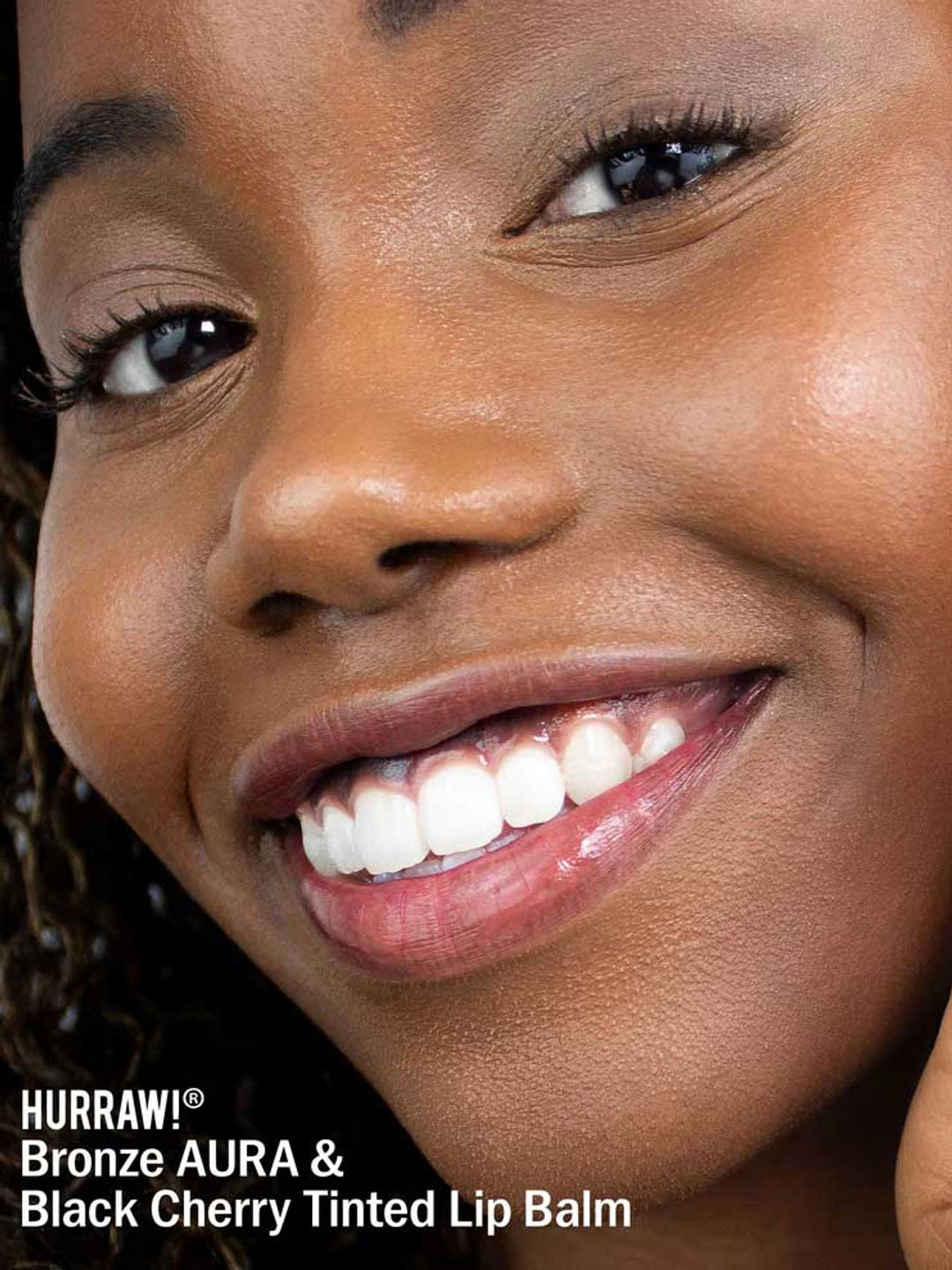 A smiling person with smooth brown skin wears a glossy shimmer on their lips. White text at the bottom reads: “Hurraw! Bronze Aura Balm 5.8g & Black Cherry Tinted Lip Balm.”.