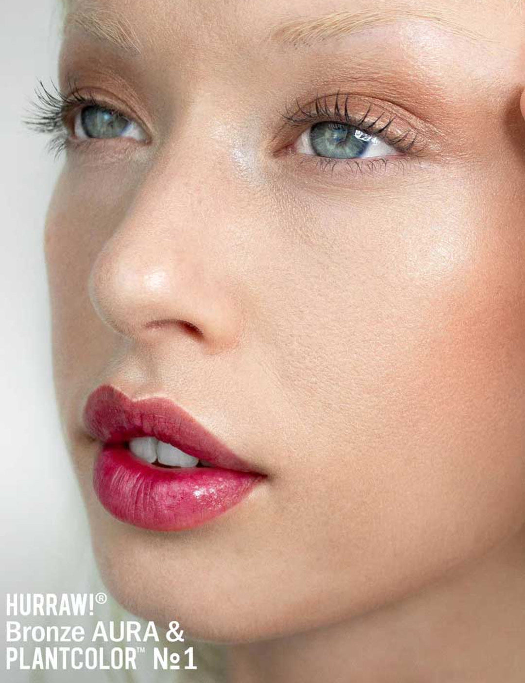 A close-up of a person’s face with dewy skin, natural makeup, shimmery bronze eyeshadow, defined lashes, and glossy pink lips enhanced by Hurraw! Bronze Aura Balm 5.8g. Text below: Hurraw! Bronze Aura & PLANTCOLOR No1.