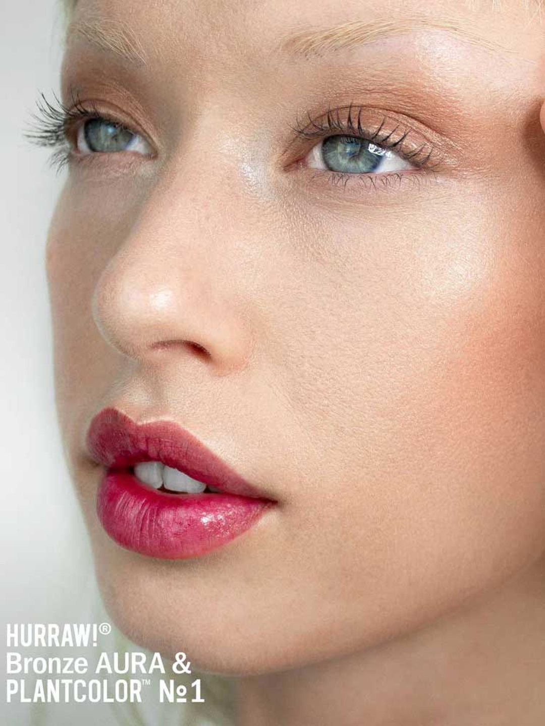 A close-up of a person’s face with dewy skin, natural makeup, shimmery bronze eyeshadow, defined lashes, and glossy pink lips enhanced by Hurraw! Bronze Aura Balm 5.8g. Text below: Hurraw! Bronze Aura & PLANTCOLOR No1.