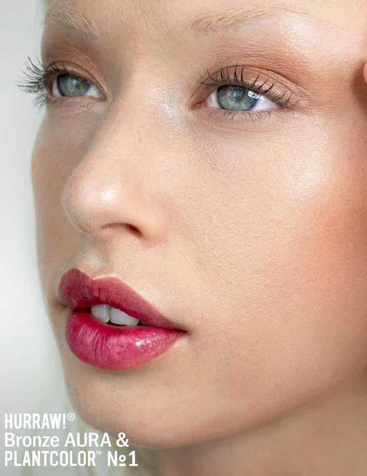 A close-up of a person’s face with dewy skin, natural makeup, shimmery bronze eyeshadow, defined lashes, and glossy pink lips enhanced by Hurraw! Bronze Aura Balm 5.8g. Text below: Hurraw! Bronze Aura & PLANTCOLOR No1.