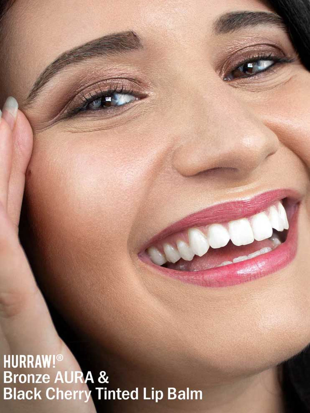 A close-up of a smiling person with glossy, tinted lips and glowing skin. Text on the image: Hurraw! Bronze Aura Balm 5.8g—your go-to bronze shimmer balm and multi-use tint for radiant looks.