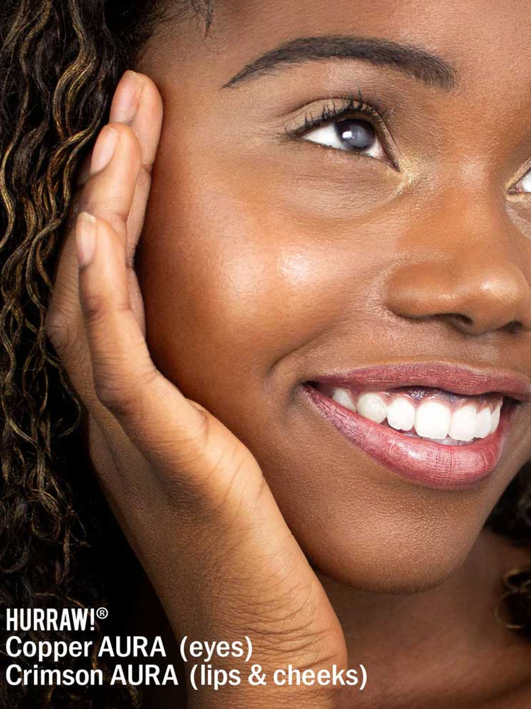 A close-up of a smiling woman with braided hair, glowing with copper eye makeup and crimson lips and cheeks. Her hand touches her face. Text below highlights Hurraw! Copper Aura Balm 5.8g, a vegan shimmer balm for multi-use radiance.