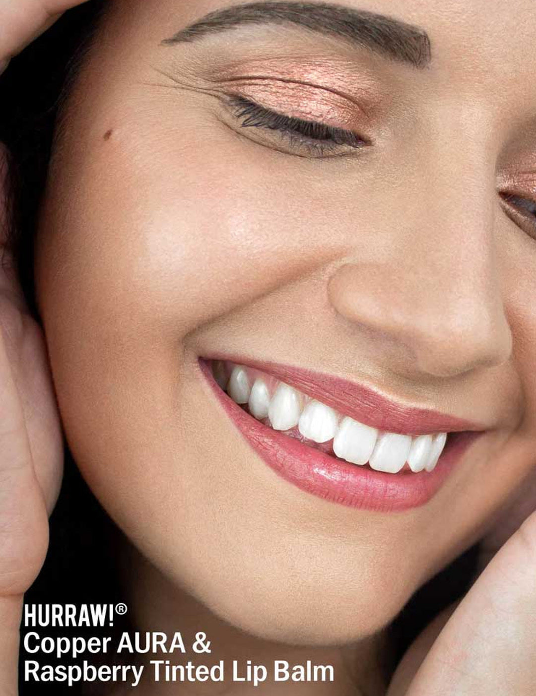 A smiling person wears shimmery copper eyeshadow and pink-tinted lips. Text: “Hurraw! Copper Aura Balm 5.8g—vegan shimmer balm and multi-use highlighter for eyes, cheeks, or lips.”.