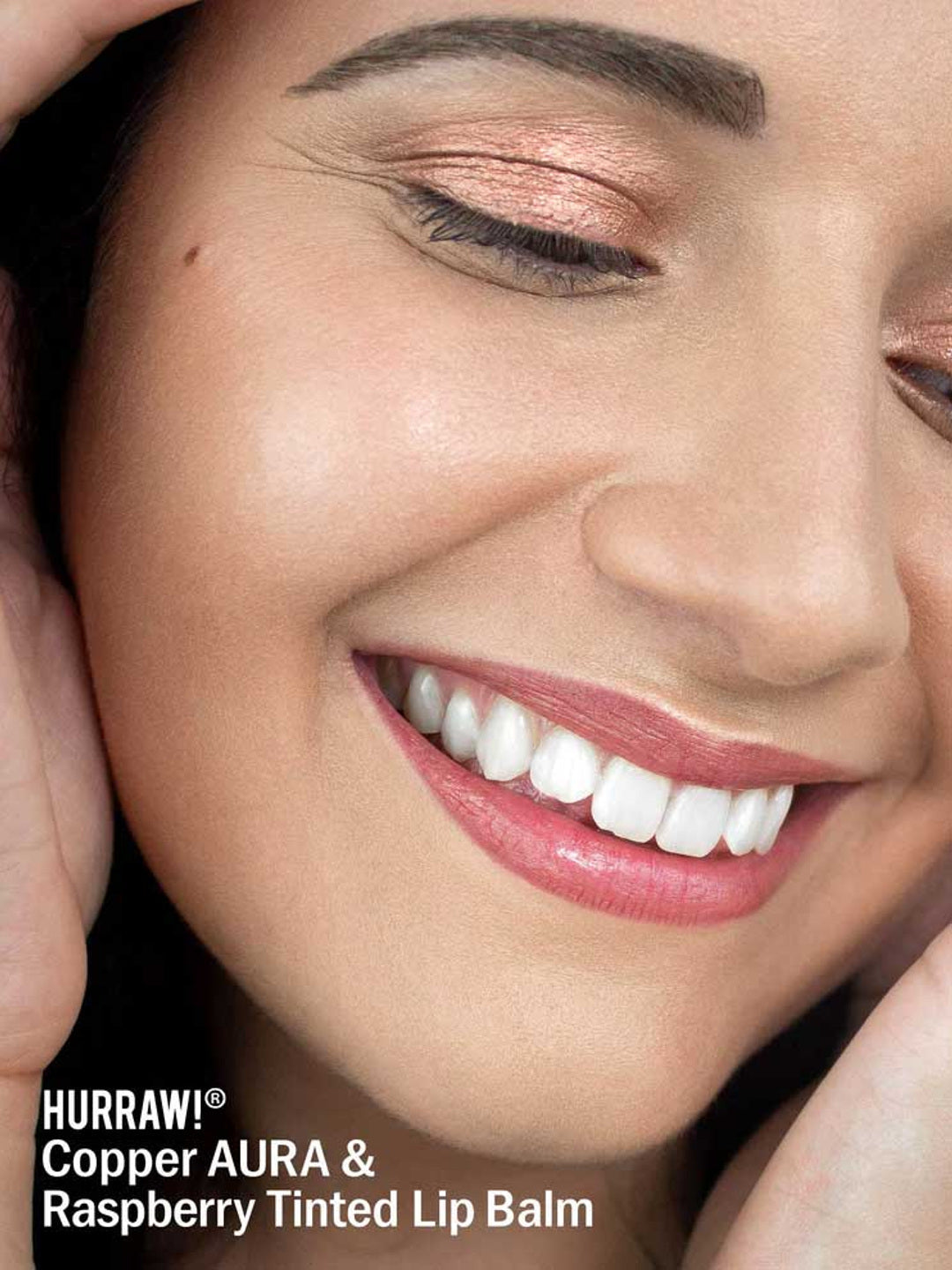 A smiling person wears shimmery copper eyeshadow and pink-tinted lips. Text: “Hurraw! Copper Aura Balm 5.8g—vegan shimmer balm and multi-use highlighter for eyes, cheeks, or lips.”.