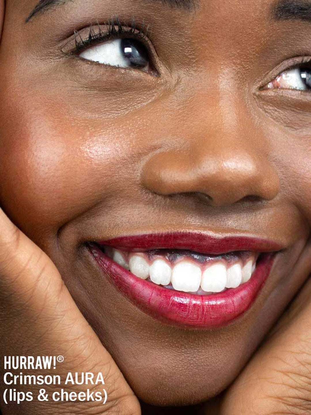 A person smiles, hands framing smooth skin and bold red lips. Text: Hurraw! Crimson Aura Balm 5.8g—vegan shimmer balm for lips & cheeks.