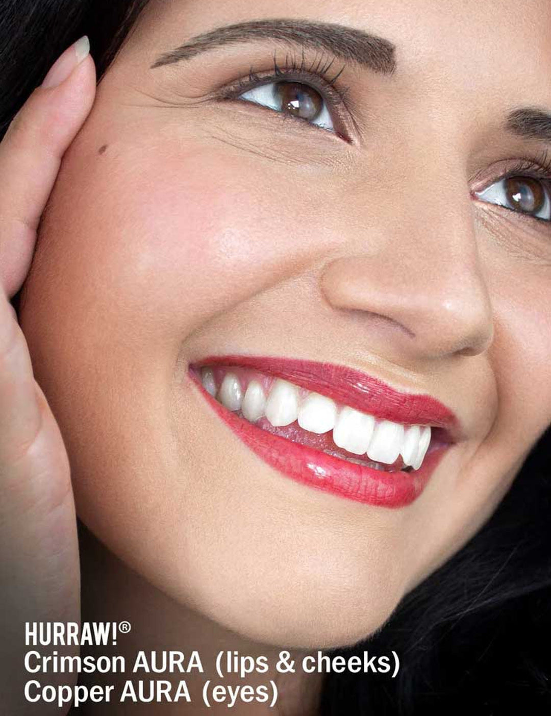 Close-up of a smiling woman with radiant skin, glossy crimson lips, rosy cheeks, and copper eyeshadow. She gently touches her face. Text below highlights Hurraw! Crimson Aura Balm 5.8g, a vegan shimmer balm and plant-based highlighter for glow.