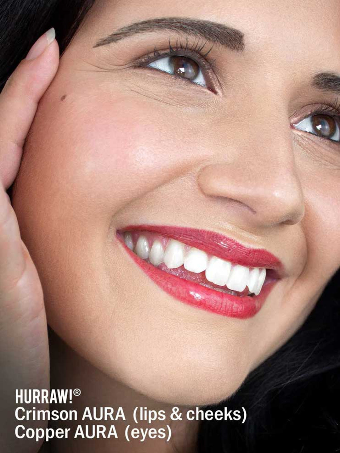 Close-up of a smiling woman with radiant skin, glossy crimson lips, rosy cheeks, and copper eyeshadow. She gently touches her face. Text below highlights Hurraw! Crimson Aura Balm 5.8g, a vegan shimmer balm and plant-based highlighter for glow.