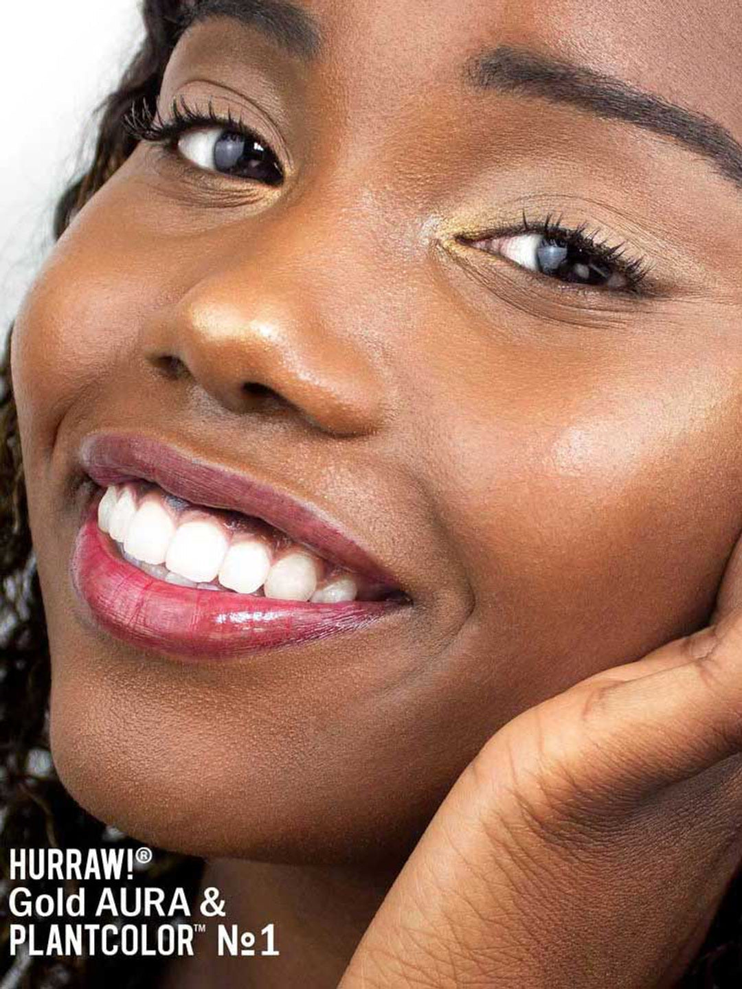 A close-up of a smiling person with radiant, glowing skin and glossy lips. Their hand gently touches their face. Text in the corner reads: “Hurraw! Gold Aura Balm 5.8g vegan highlighter.”.