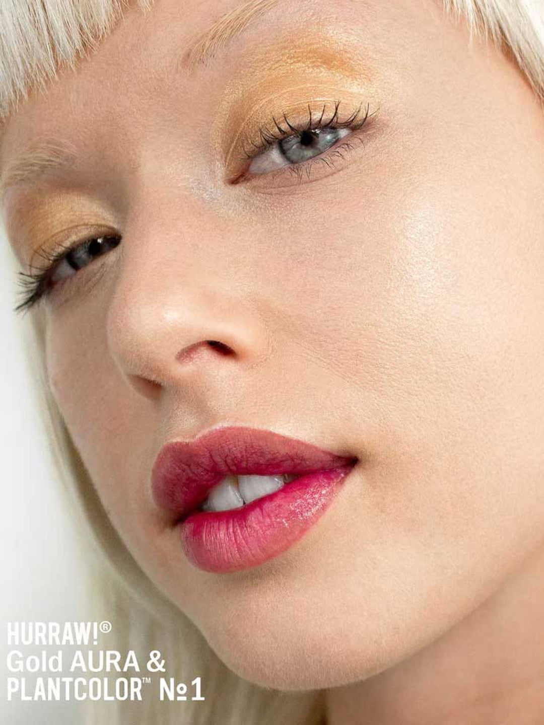 Close-up of a fair-skinned, blonde-haired person with gold eyeshadow, long lashes, and pink lipstick. Text reads: Hurraw! Gold Aura Balm 5.8g—featuring our vegan highlighter for a luminous glow.