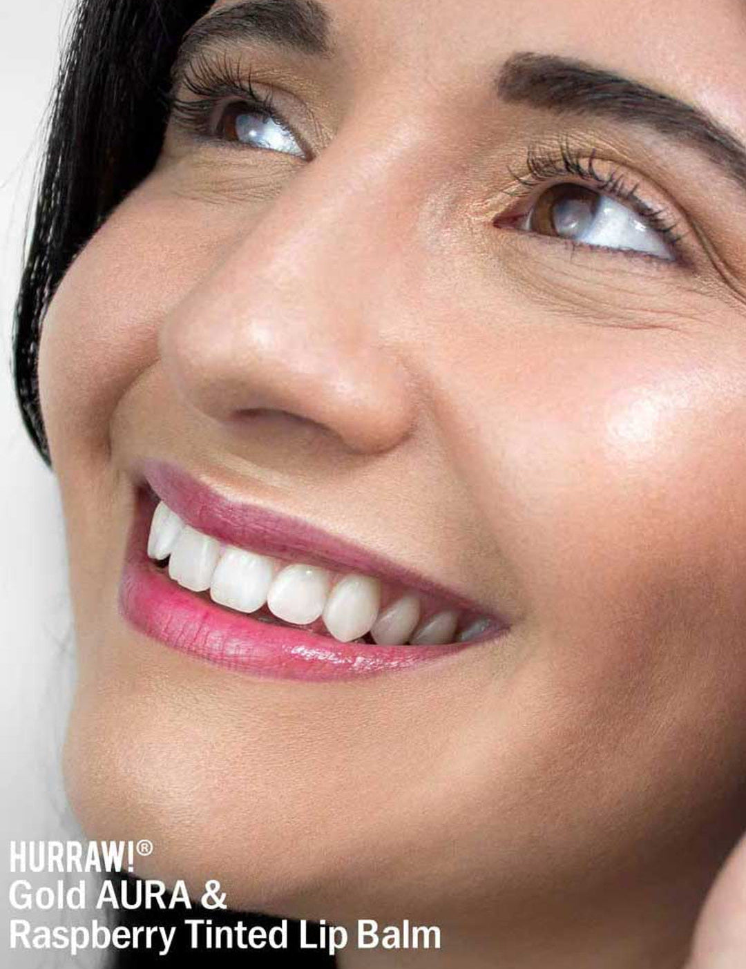 A smiling person with smooth skin and glossy, raspberry-tinted lips looks upward. Text at the lower left reads: Hurraw! Gold Aura Balm 5.8g—vegan highlighter and multi-use shimmer balm.