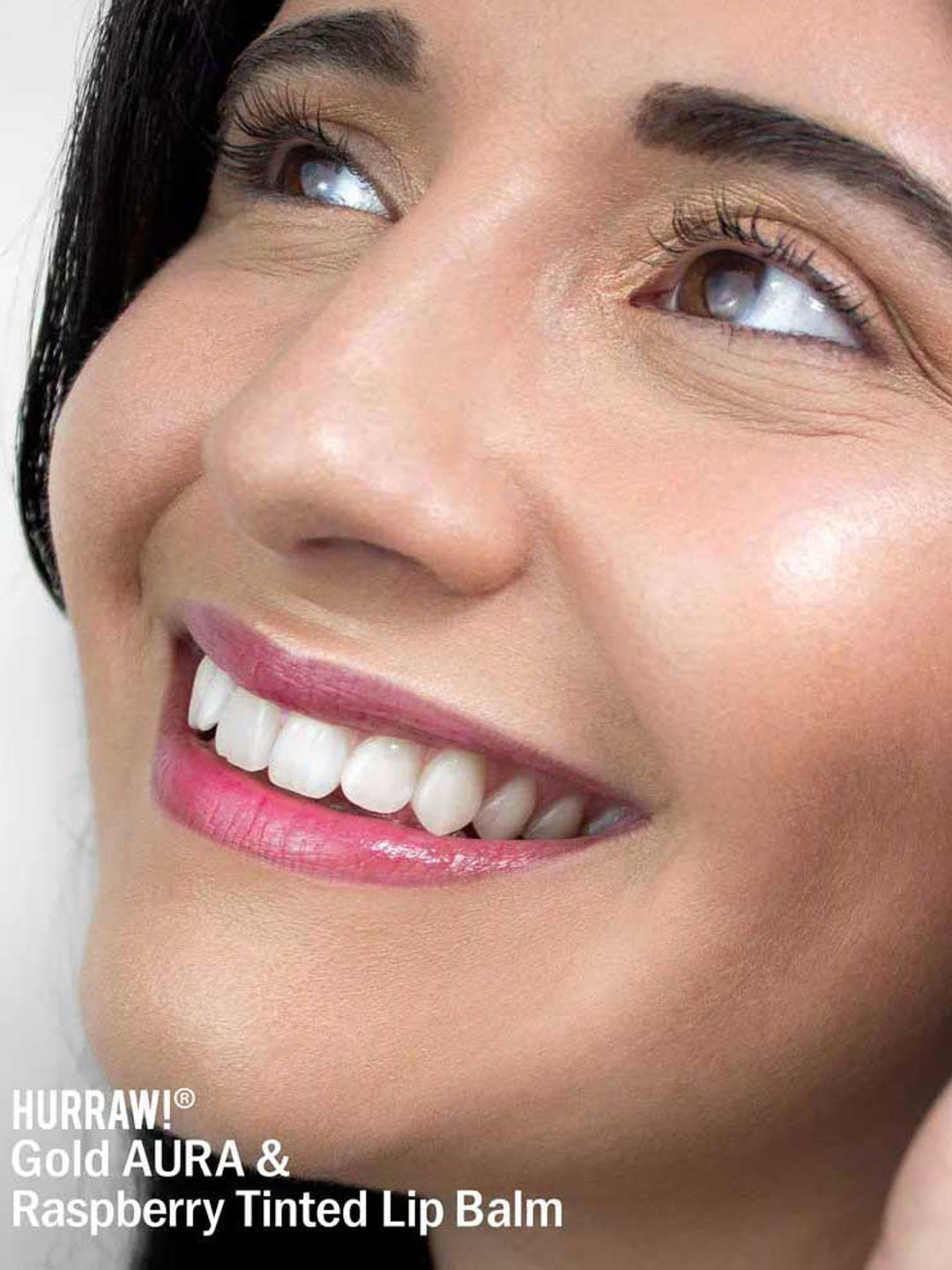 A smiling person with smooth skin and glossy, raspberry-tinted lips looks upward. Text at the lower left reads: Hurraw! Gold Aura Balm 5.8g—vegan highlighter and multi-use shimmer balm.