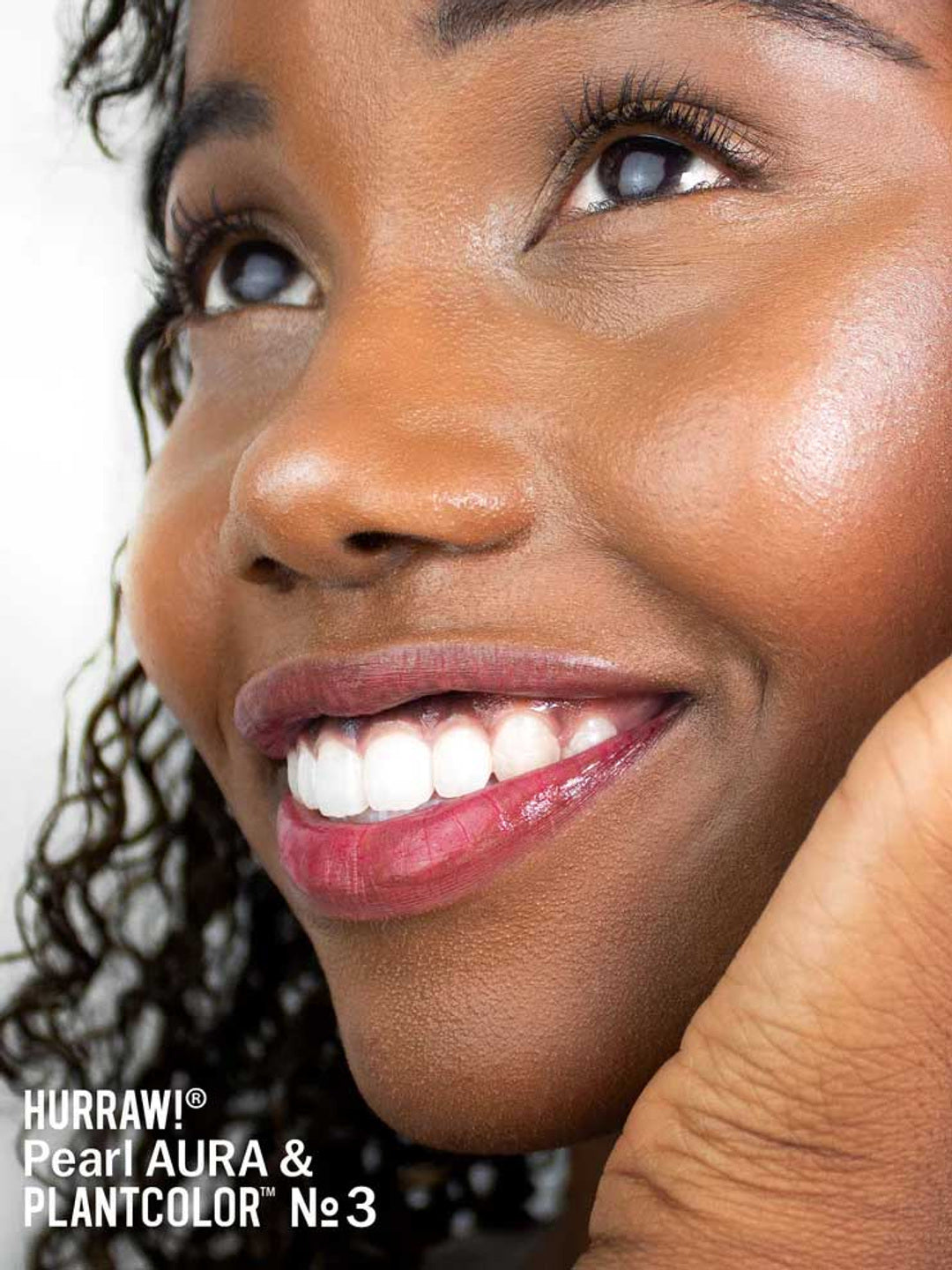 A close-up of a smiling person with glowing brown skin, curly hair, and glossy lips. Text reads: Hurraw! Pearl Aura Balm 5.8g vegan highlighter in the lower left corner.