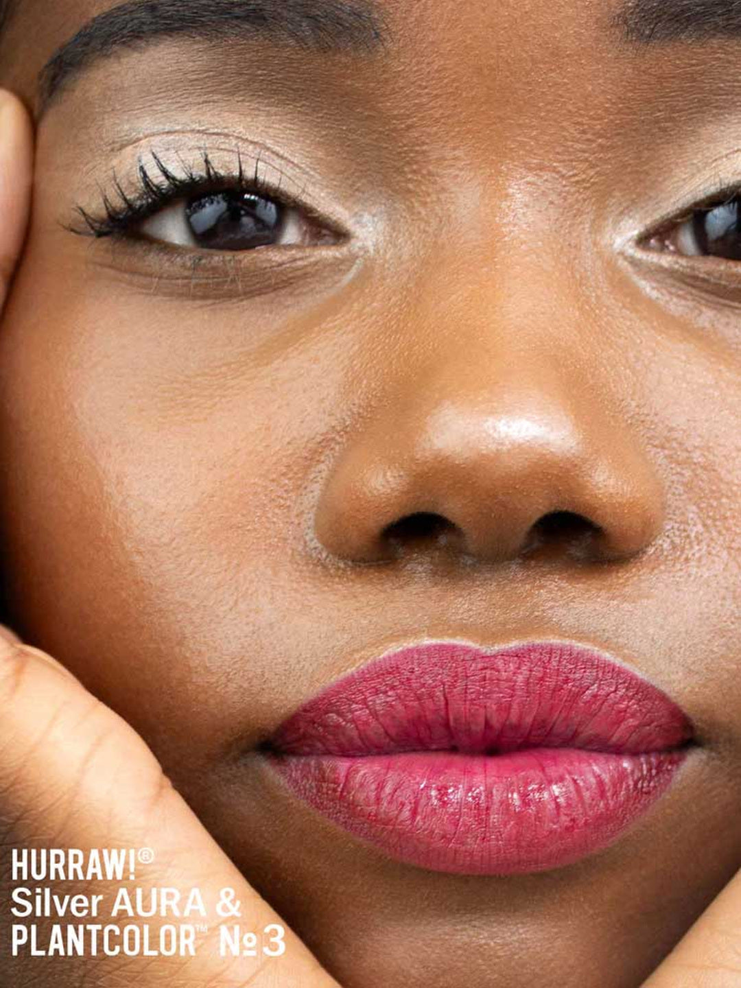 Close-up of a person with smooth dark skin, natural makeup, bold pinkish-red lips, and hands on cheeks. Text: Hurraw! Silver Aura Balm vegan highlighter & PLANTCOLOR No. 3.