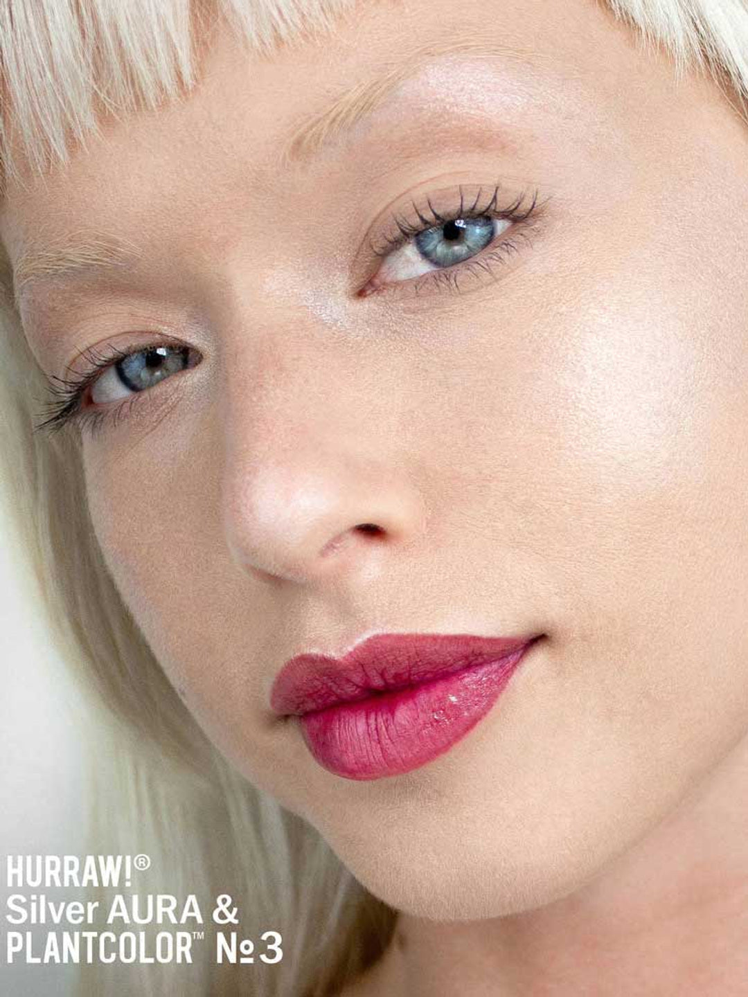 Close-up of a person with platinum blonde hair and glowing skin, wearing bold pink lipstick. The text “Hurraw! Silver Aura Balm” appears in the bottom left corner, highlighting the vegan silver shimmer highlighter.