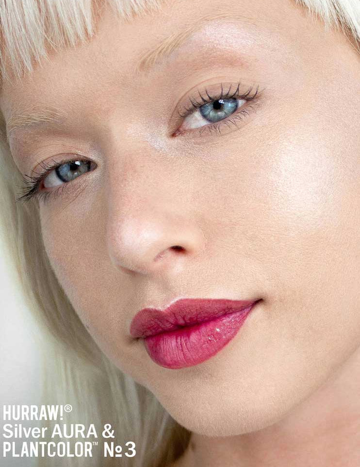 Close-up of a person with platinum blonde hair and glowing skin, wearing bold pink lipstick. The text “Hurraw! Silver Aura Balm” appears in the bottom left corner, highlighting the vegan silver shimmer highlighter.