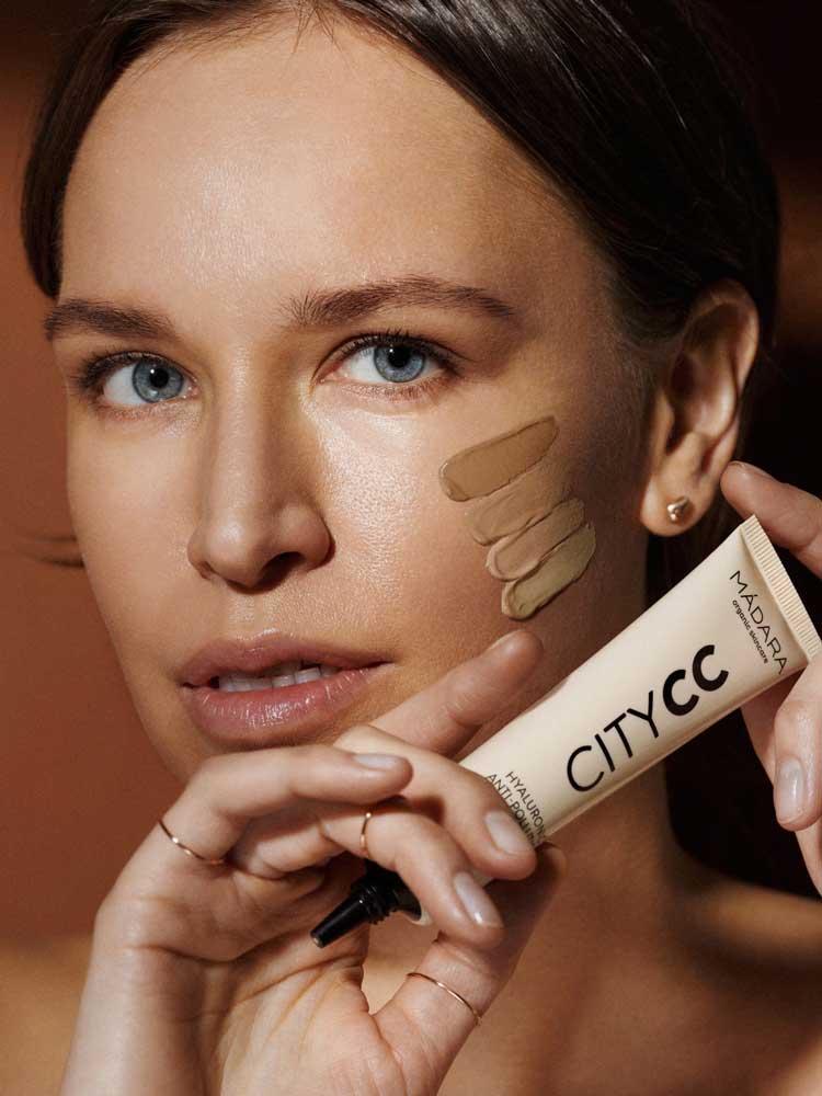 A woman with clear skin and blue eyes holds a tube of Hyaluronic Anti-Pollution CC Cream SPF15 40ml near her face, displaying three foundation shades on her cheek to showcase its coverage and protective skincare benefits.
