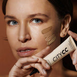 A woman with clear skin and blue eyes holds a tube of Hyaluronic Anti-Pollution CC Cream SPF15 40ml near her face, displaying three foundation shades on her cheek to showcase its coverage and protective skincare benefits.