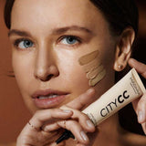 A woman with clear skin and blue eyes holds a tube of Hyaluronic Anti-Pollution CC Cream SPF15 40ml near her face, displaying three foundation shades on her cheek to showcase its coverage and protective skincare benefits.