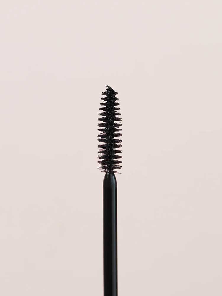 A close-up of the INIKA Organic Bold Lash Mascara Black 13ml wand, its bristles coated in black mascara, is shown vertically against a plain light beige background.