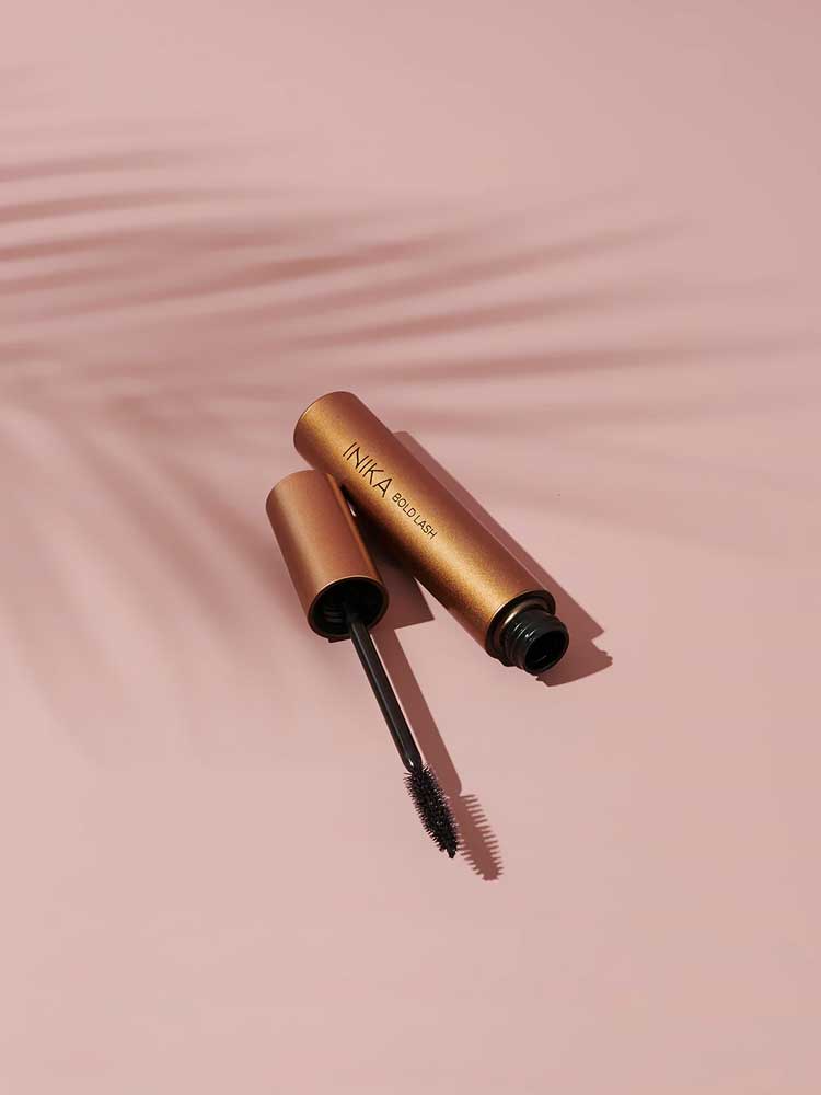 An open tube of INIKA Organic Bold Lash Mascara Black 13ml, with the wand in front, lies on a pink surface. A faint palm leaf shadow appears in the background.