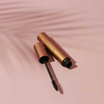 An open tube of INIKA Organic Bold Lash Mascara Black 13ml, with the wand in front, lies on a pink surface. A faint palm leaf shadow appears in the background.