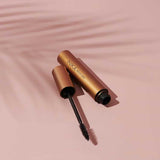 An open tube of INIKA Organic Bold Lash Mascara Black 13ml, with the wand in front, lies on a pink surface. A faint palm leaf shadow appears in the background.