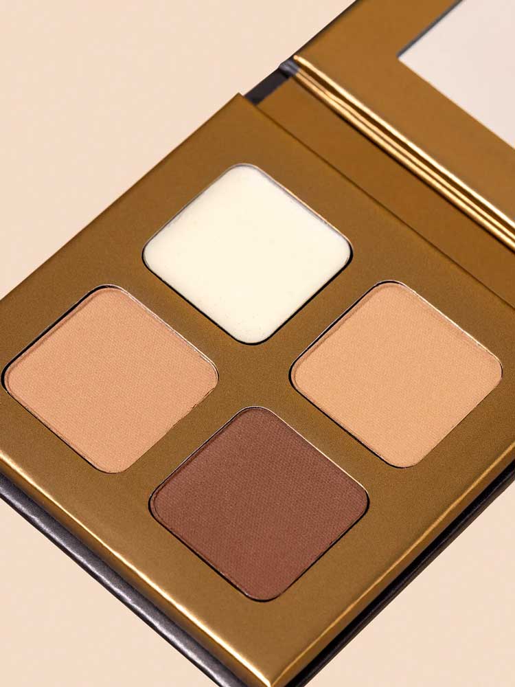 A close-up of the INIKA Organic Brow Palette 8g, an open vegan set with four shades—off-white, light beige, medium tan, and deep brown—in a gold case against a light background.