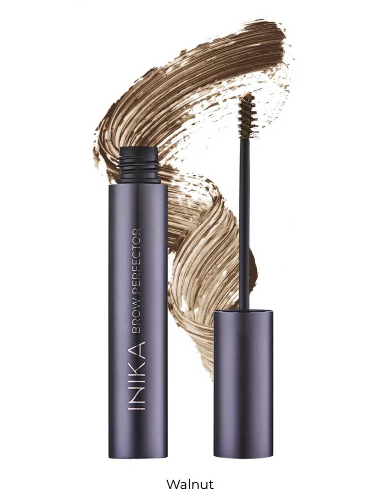 A tube of INIKA Organic Brow Perfector 6.5ml in Walnut stands open with its brush visible, set against a background of a natural eyebrow gel smear.