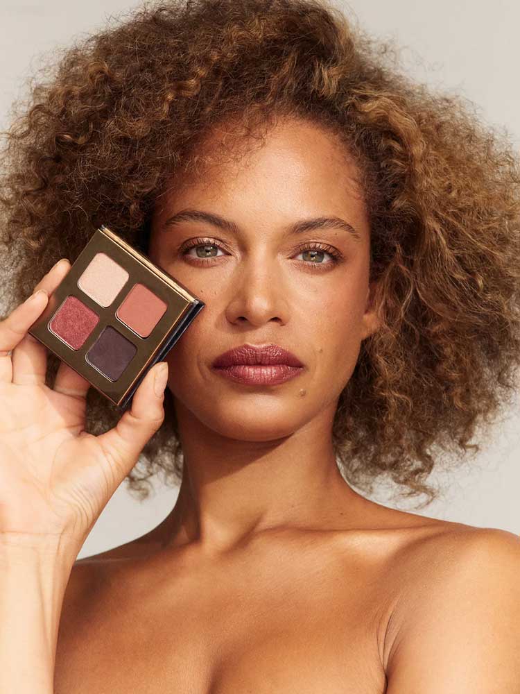 A woman with curly brown hair holds the INIKA Organic Eyeshadow Quad - Flowers 8g, featuring four vegan shades, next to her face. She wears natural makeup and has a neutral expression against a plain background.