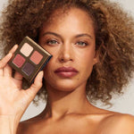A woman with curly brown hair holds the INIKA Organic Eyeshadow Quad - Flowers 8g, featuring four vegan shades, next to her face. She wears natural makeup and has a neutral expression against a plain background.