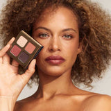A woman with curly brown hair holds the INIKA Organic Eyeshadow Quad - Flowers 8g, featuring four vegan shades, next to her face. She wears natural makeup and has a neutral expression against a plain background.