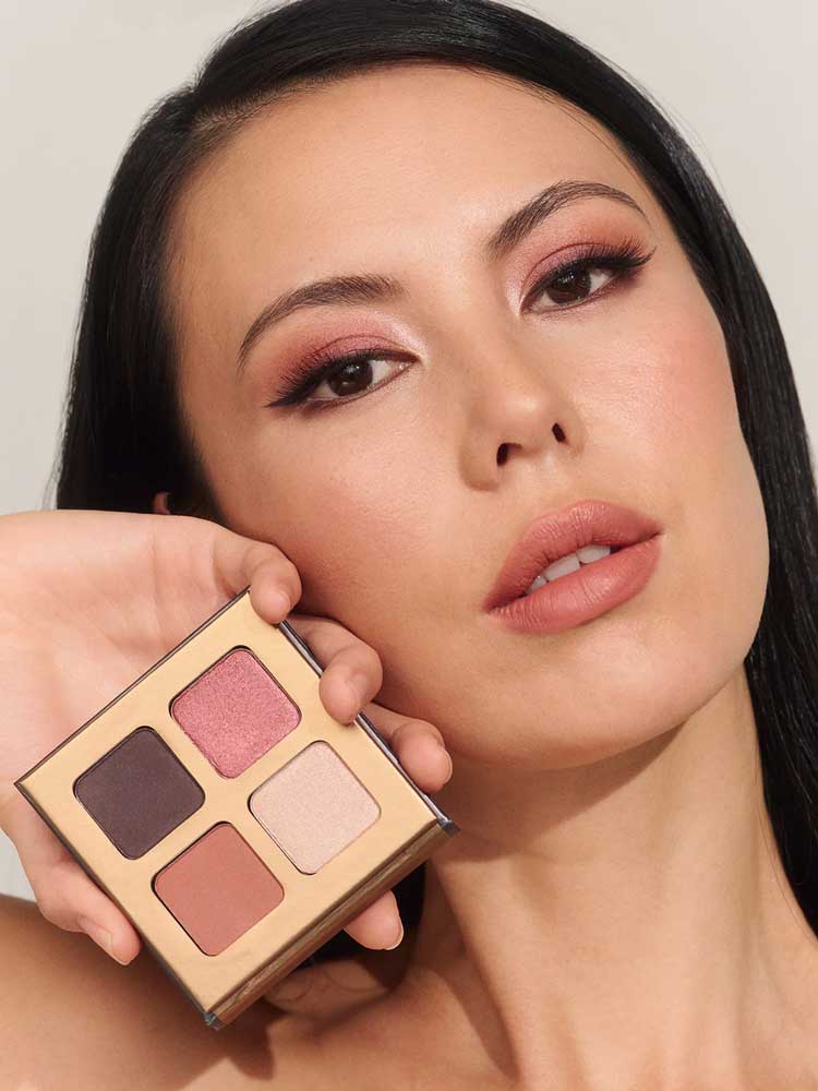 A woman with long dark hair and natural makeup holds the INIKA Organic Eyeshadow Quad - Flowers 8g near her face, displaying its four vegan shades—deep brown, rose, beige, and terracotta—and her eye makeup.