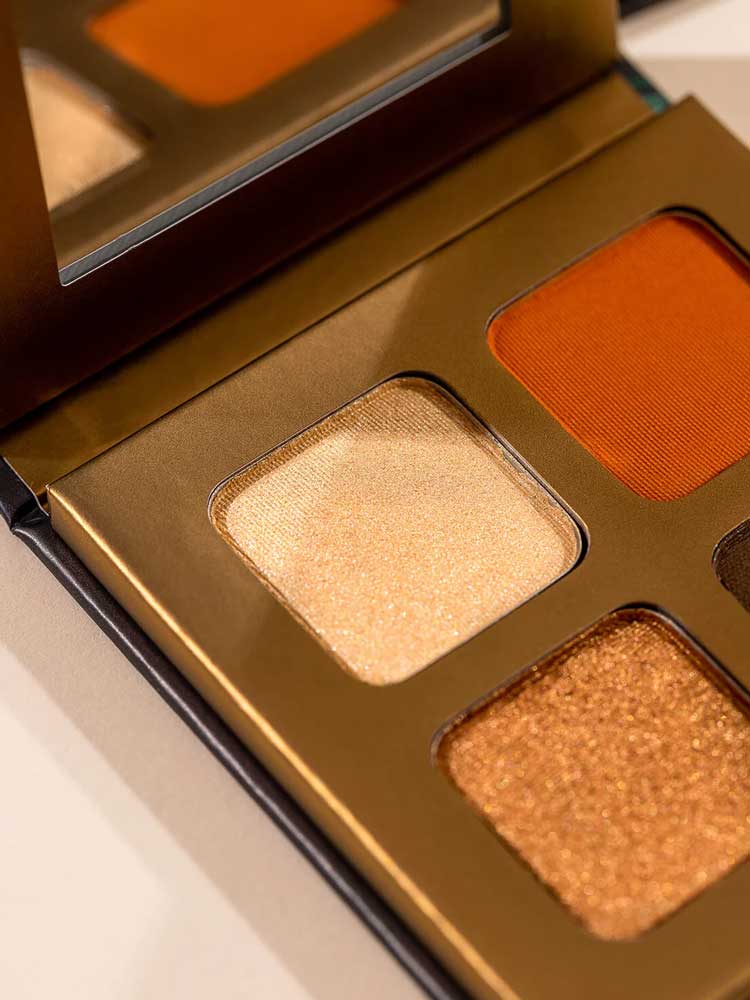 Close-up of the INIKA Organic Eyeshadow Quad - Sunset 8g, a natural palette with a mirror, featuring shimmery gold, bronze, and matte orange mineral shades in a sleek metallic gold case.
