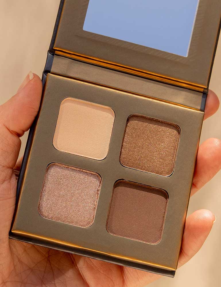 A hand holds the INIKA Organic Eyeshadow Quad - Wind 8g, featuring four neutral shades—beige, shimmery light brown, matte brown, and shimmery taupe—with a small mirror inside the lid.