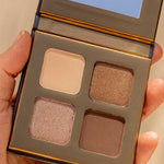 A hand holds the INIKA Organic Eyeshadow Quad - Wind 8g, featuring four neutral shades—beige, shimmery light brown, matte brown, and shimmery taupe—with a small mirror inside the lid.