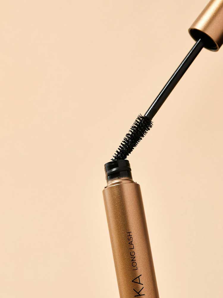 The INIKA Organic Long Lash Mascara 8ml, in a gold tube with a black wand and bristled brush partly removed from the container, is set against a beige background.
